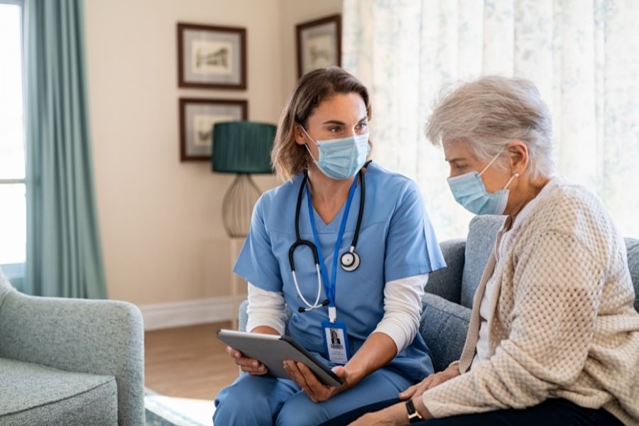 Nurse and senior woman going through medical record on digital tablet during home visit and wearing face mask. Doctor wearing protective face mask during covid pandemic and showing age friendly health systems